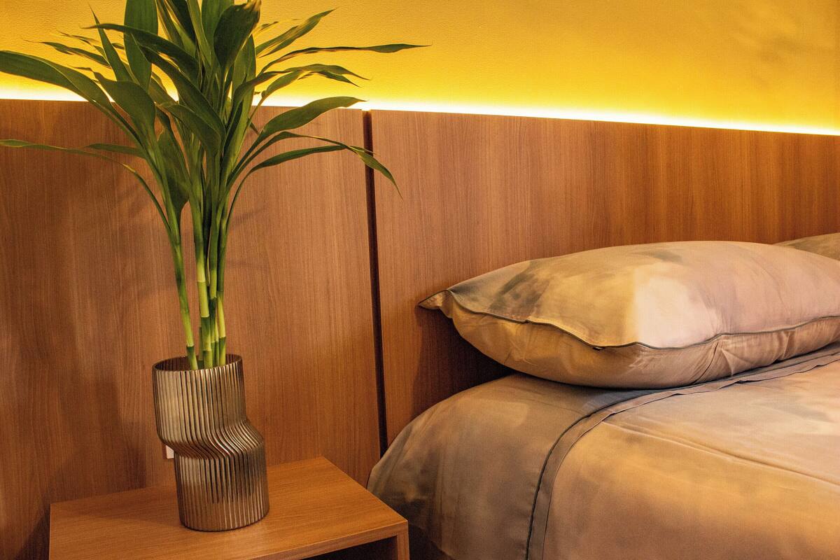 A wooden bedside table with a plant in a textured vase, a bed with a beige comforter and pillows, and a wooden headboard with a warm light underneath.