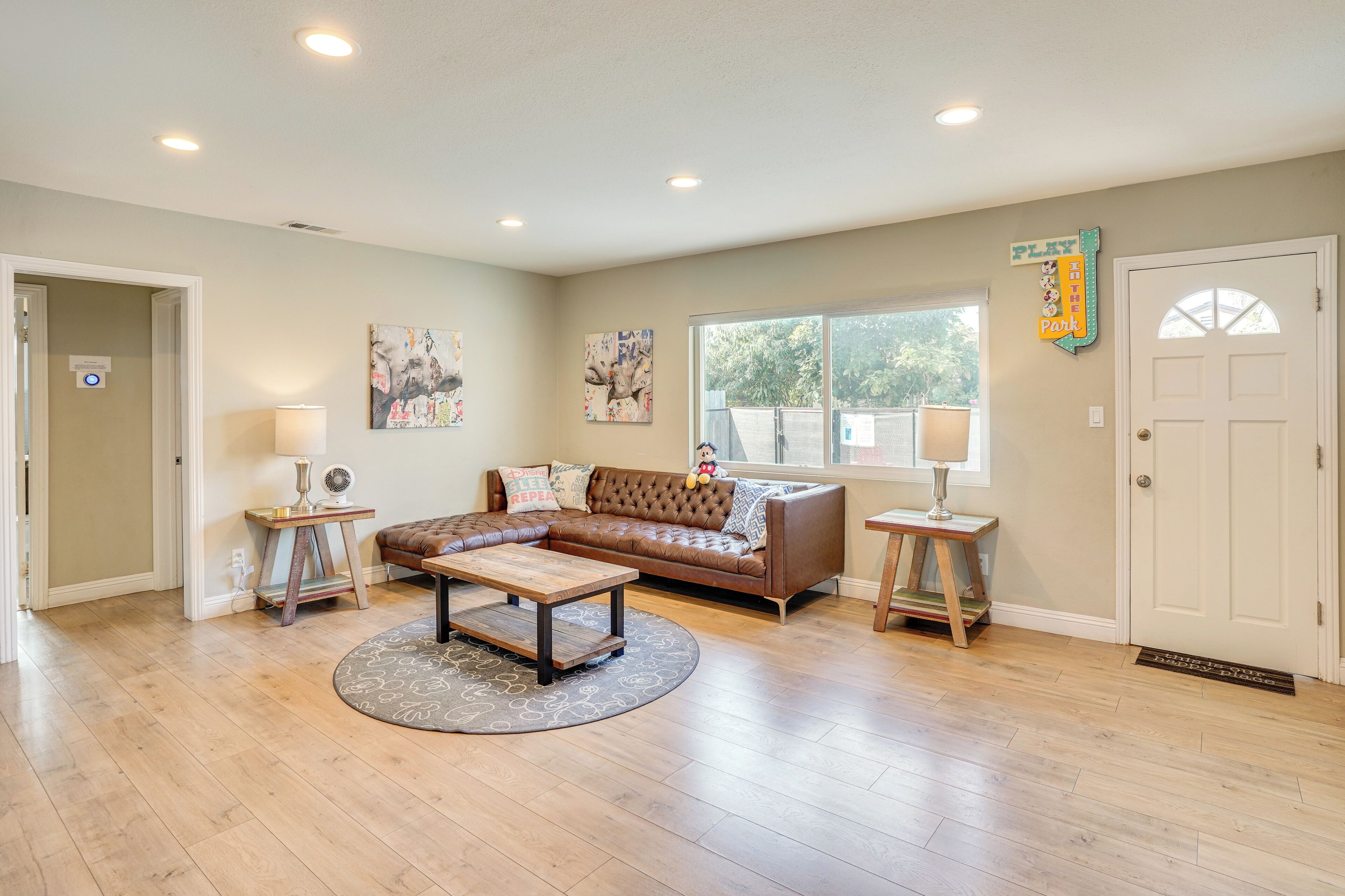 A spacious living room with a brown leather sectional sofa, a wooden coffee table, and a round rug. There are two side tables with lamps, a large window with a view of trees, and a door with a 'PLAY PARK' sign.