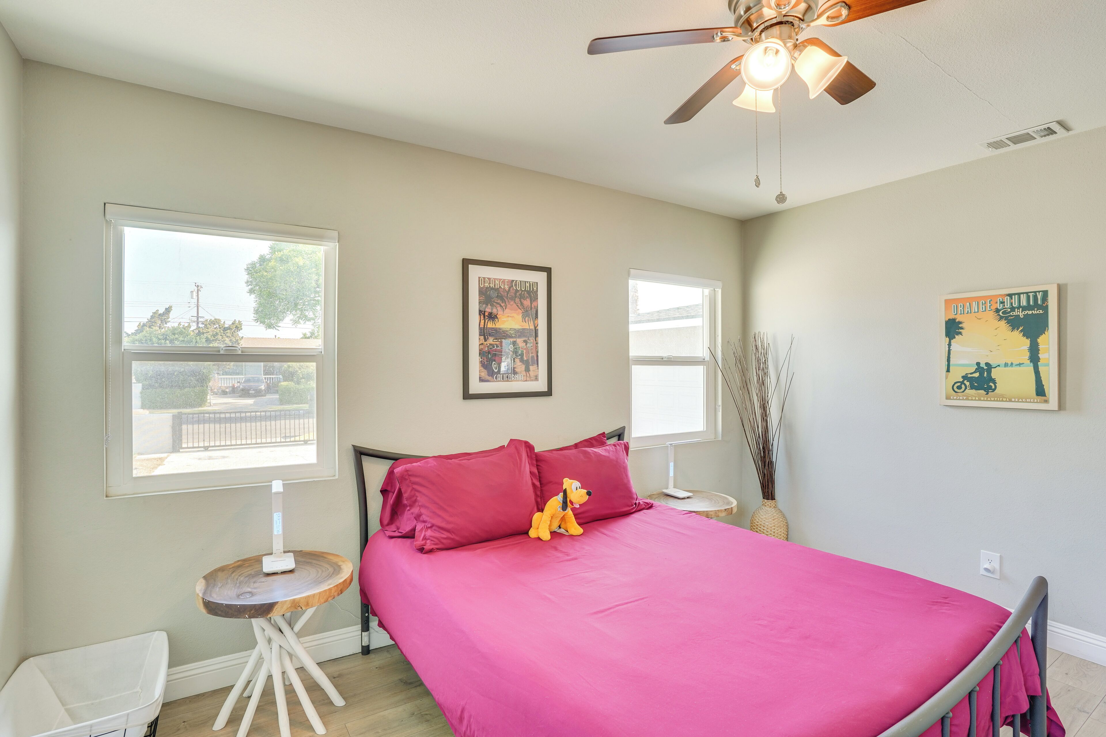 A bedroom with a bed, a ceiling fan, two windows, and a small table with a lamp.