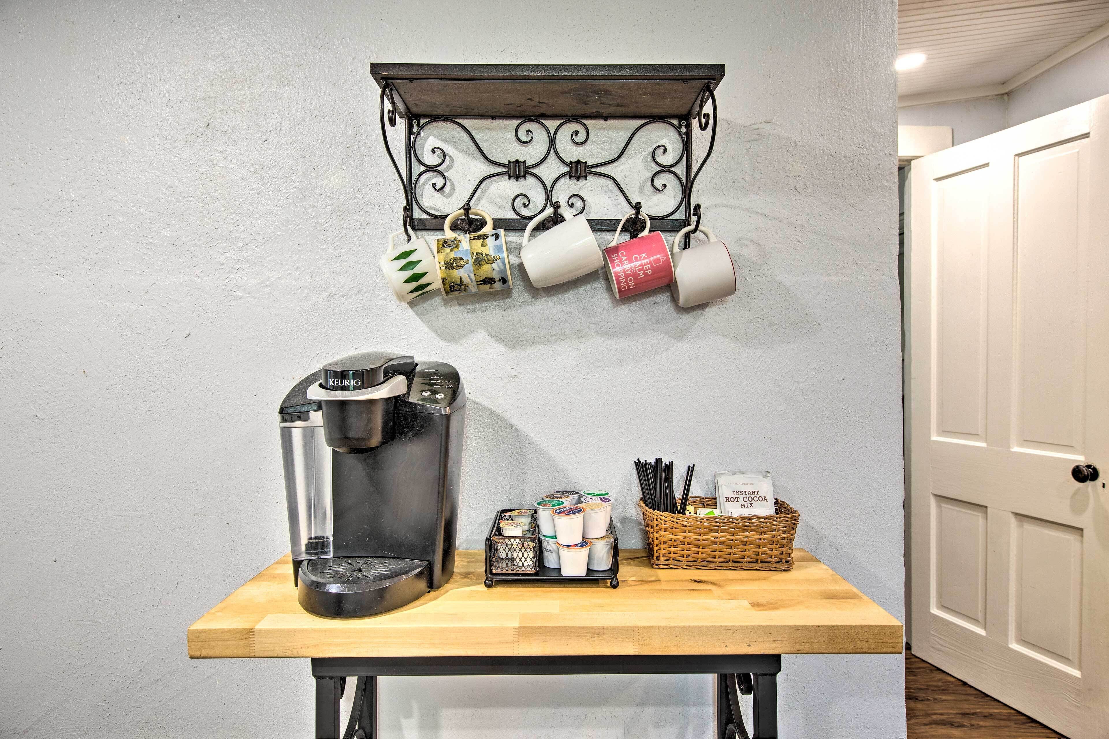 A coffee maker on a wooden table with a coffee mug, a basket of utensils, and a spice rack with containers.