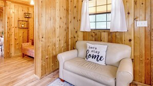 Una habitación acogedora con paredes de madera, un sofá color beige, una ventana con cortinas y una lámpara fijada a la pared.