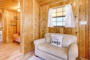 A cozy room with wooden walls, a beige sofa, a window with curtains, and a wall-mounted light fixture. - Couples Cabin w/ Luxury Deck, 1 Mi to Canyon Lake! (Canyon Lake)