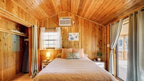 Habitación de madera con cama, ventana con cortinas, aire acondicionado en la pared y una pequeña mesa con lámpara.