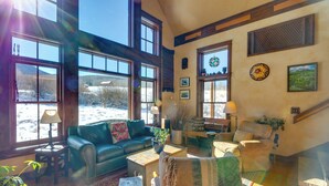 A cozy living room with a green sofa, a wooden coffee table, and a patterned rug. There are large windows with a view of a snowy landscape. - Solar-powered Home w/ Mtn Views 33 Mi to Telluride (Placerville)