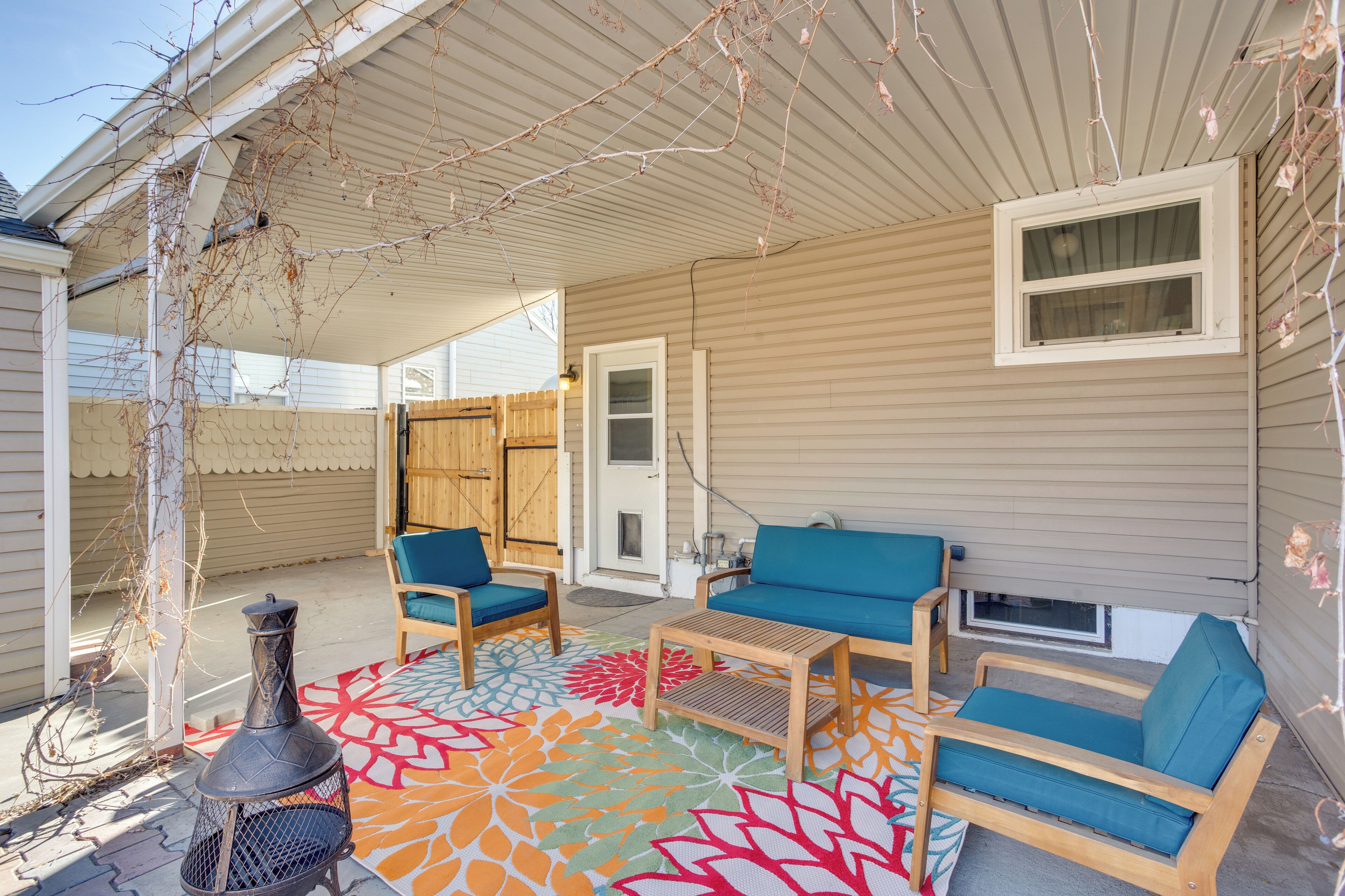 A covered patio with a fire pit, two blue cushioned chairs, a wooden table, and a colorful rug.