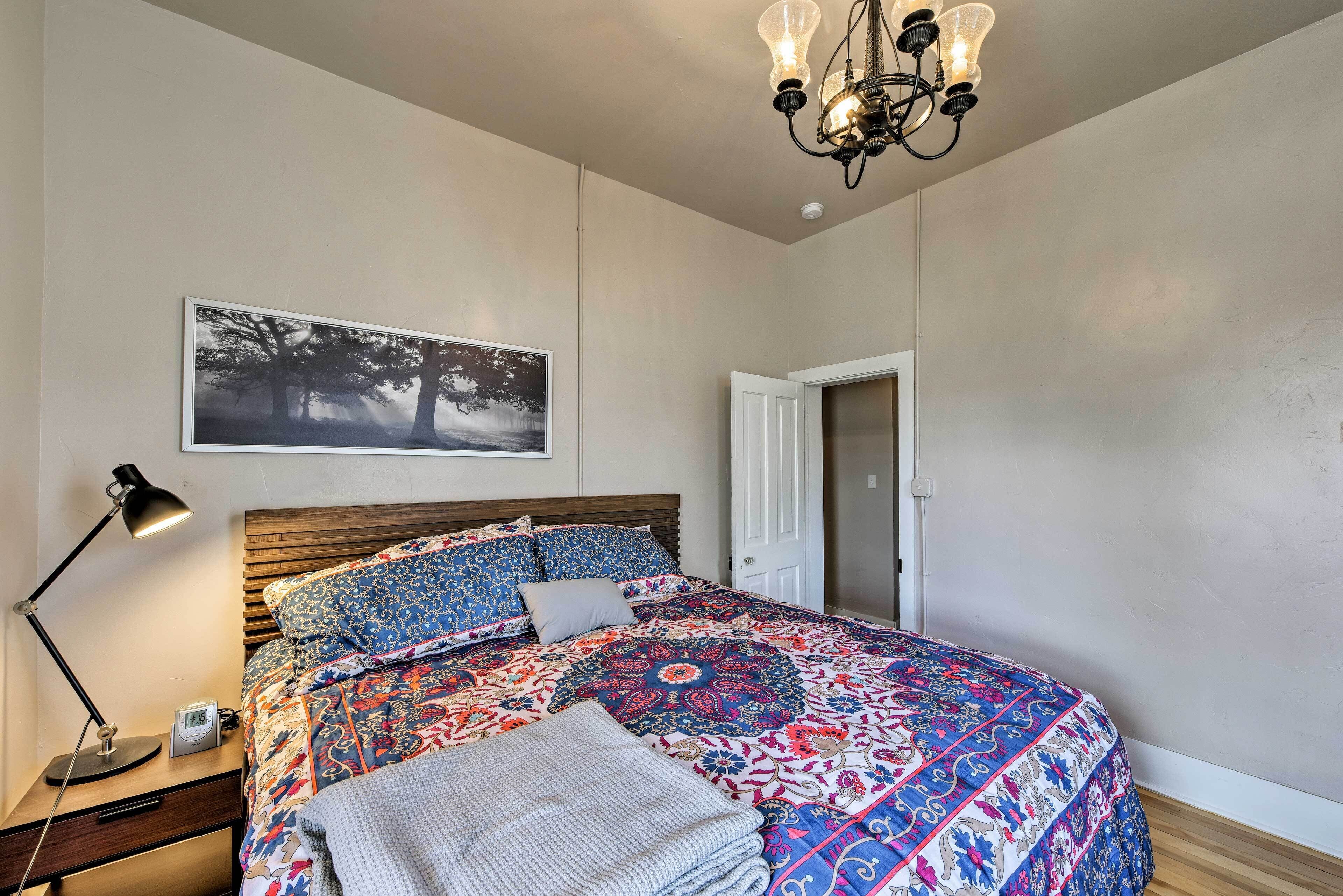 A bedroom with a large bed, a nightstand with a lamp, a framed picture on the wall, and a door.