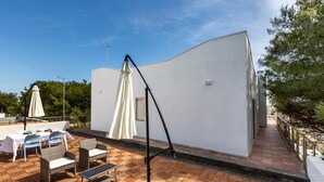 Um pátio moderno ao ar livre, com parede branca, piso de madeira e mobiliário externo, incluindo cadeiras e mesas.
