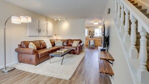 Una sala moderna con un sofá seccional de cuero marrón, un tapete blanco y una mesa de centro de madera. También se ve una lámpara de pie y un televisor.