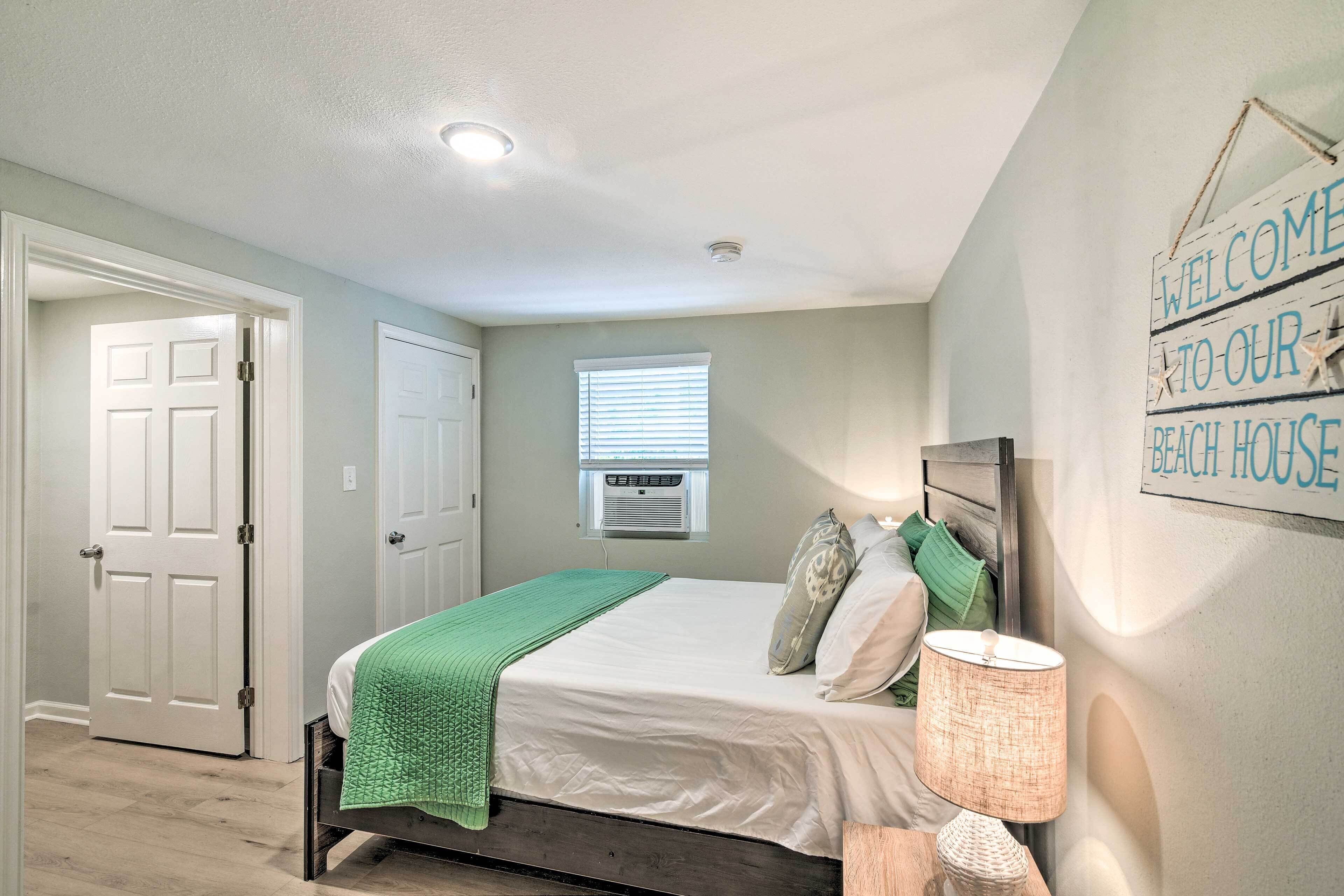 A bedroom with a bed, a green throw blanket, a lamp, and a 'Welcome to our Beach House' sign.