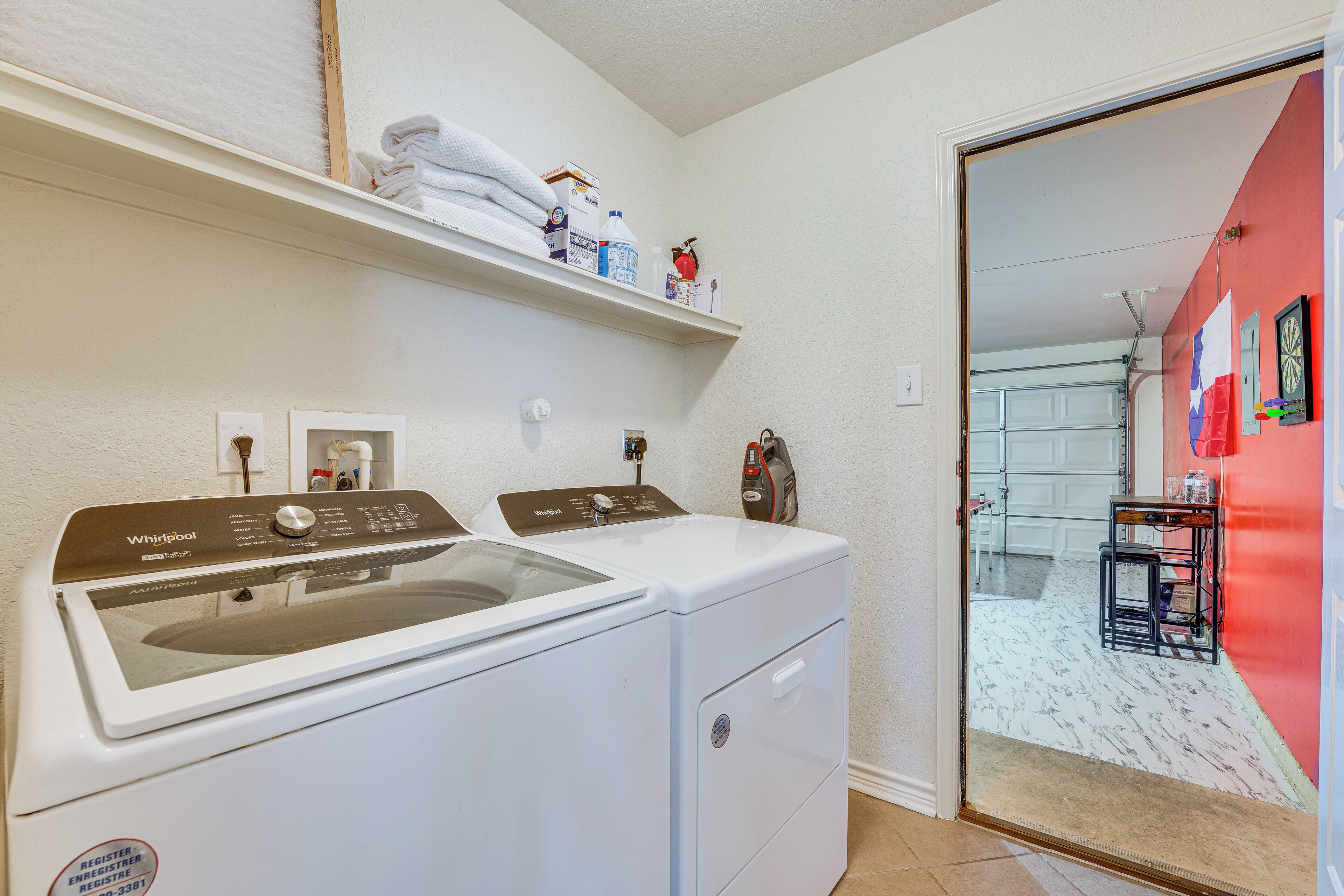 A laundry area with a Whirlpool washer and dryer, a shelf with folded towels and laundry supplies, and a red wall with a mirror and decorations.