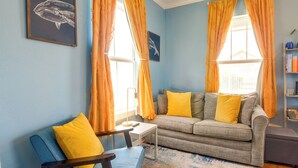 Ein Wohnzimmer mit Sofa, Sessel und Couchtisch. Zwei Fenster mit Vorhängen, eine Wand mit einem gerahmten Bild und eine Bücherwand mit Büchern.