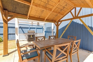 An outdoor patio with a wooden table and chairs, a grill, and a blue fence in the background.