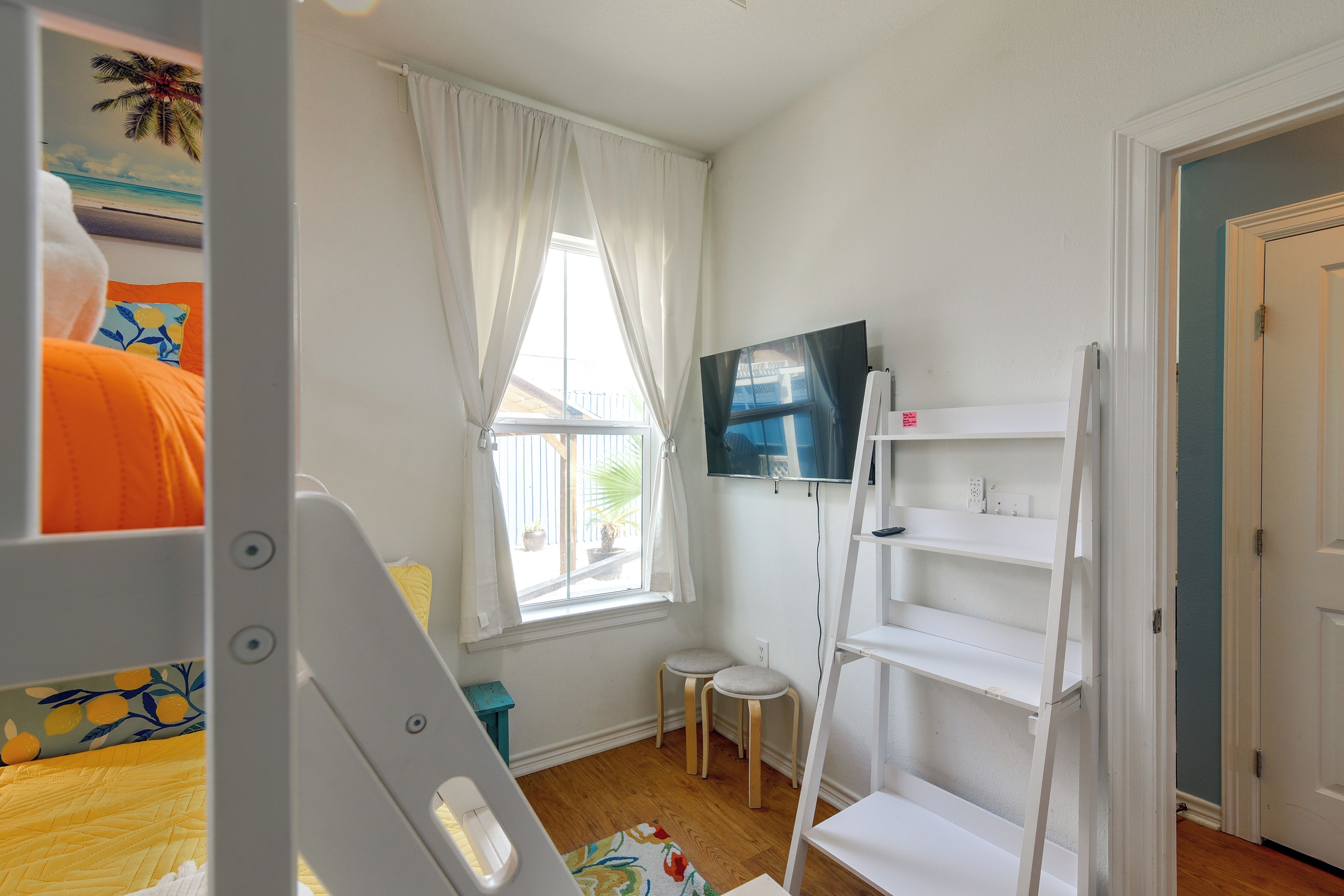 Une chambre d’enfant avec un lit superposé, un petit tabouret, un téléviseur posé sur un meuble et une fenêtre avec des rideaux.