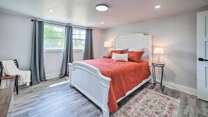 A bedroom with a white bed, grey curtains, a small table with a lamp, and a wooden floor.