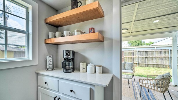 A kitchen with white cabinets, wooden shelves holding mugs and a coffee maker, and a view of a backyard with a chair and a table.