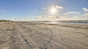 A sandy beach with footprints and tire tracks, leading to the ocean under a clear sky with the sun shining. - Ocean-view Beach House in Galveston, Dogs Welcome! (Galveston)