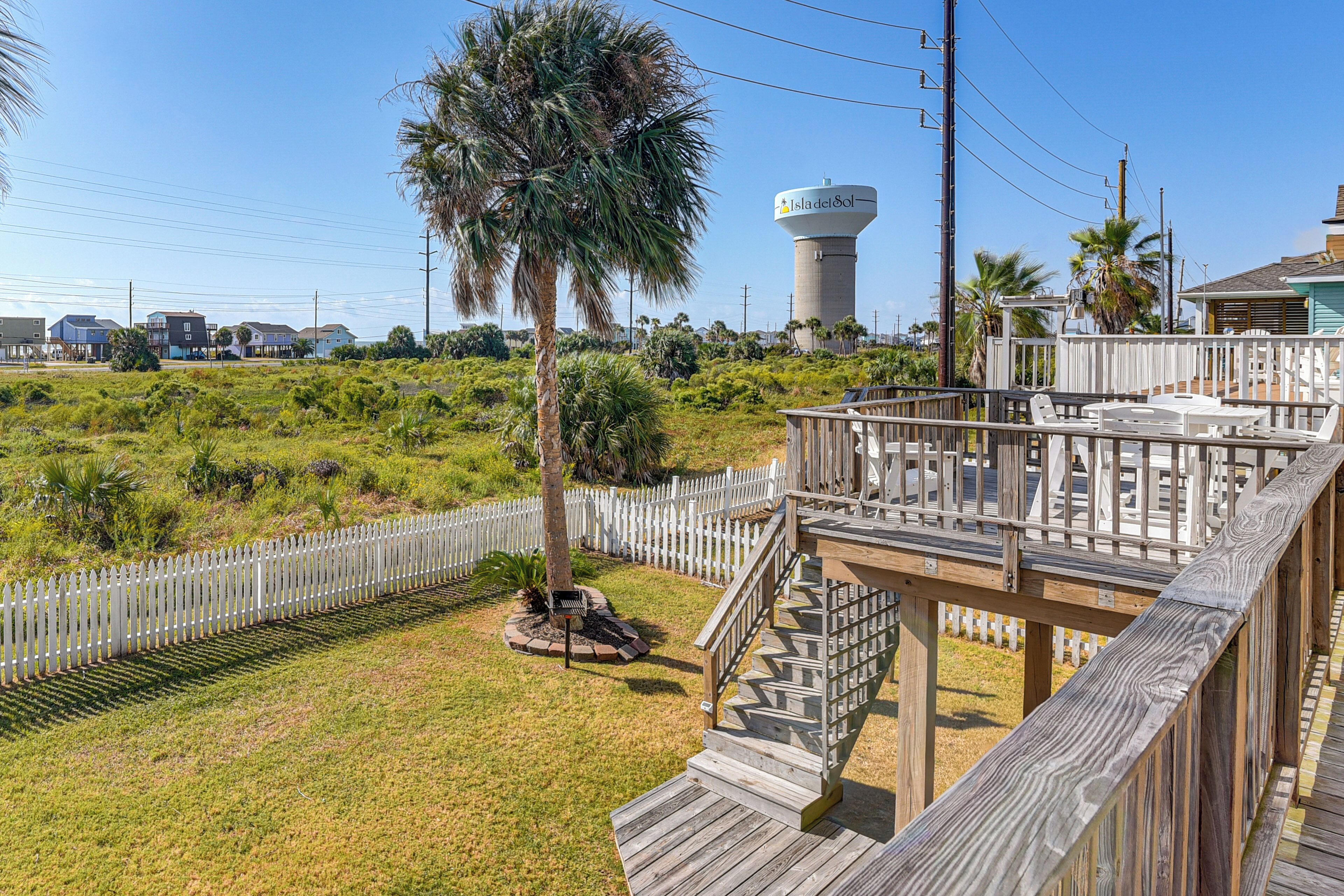 A wooden deck with a staircase leading to a covered area, surrounded by a white picket fence and lush greenery.