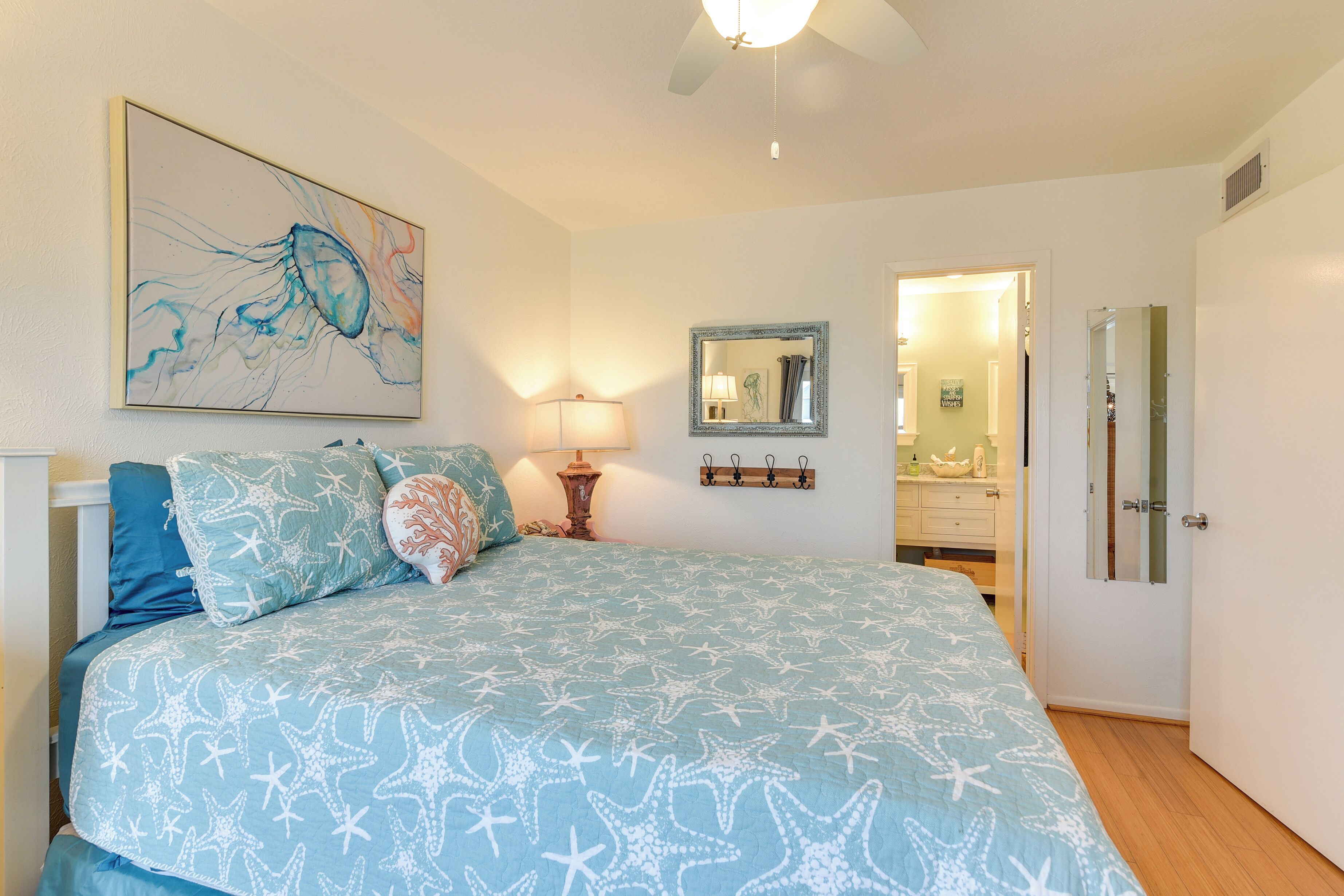 A bedroom with a bed featuring a starfish pattern, a nightstand with a lamp, a wall-mounted mirror, and a painting of a jellyfish.
