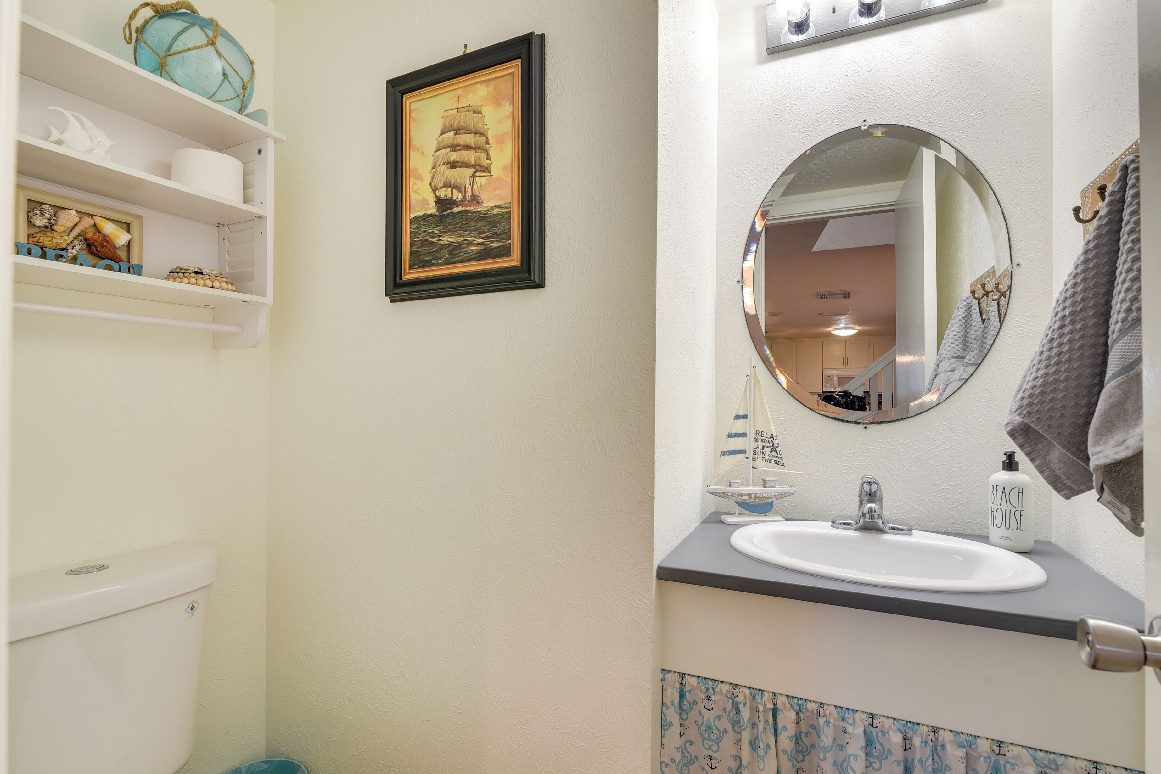 A bathroom with a toilet, a sink with a mirror, shelves with decorative items, and a framed picture of a ship.