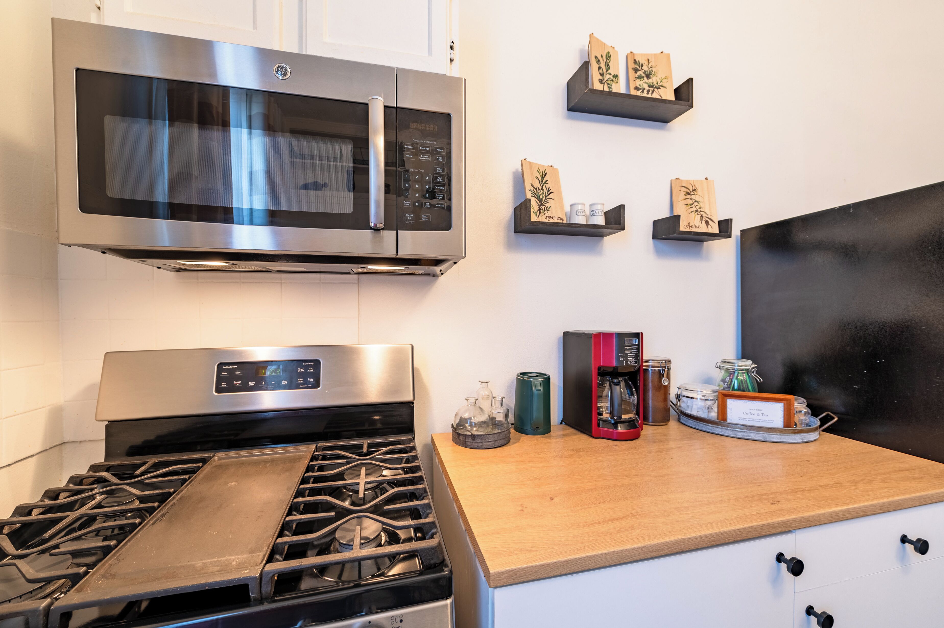 A kitchen with a stainless steel microwave, a gas stove, and wooden shelves with jars and bags.