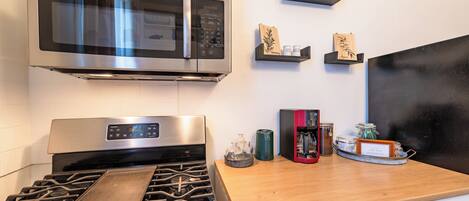A kitchen with a stainless steel microwave, a gas stove, and wooden shelves with jars and bags.
