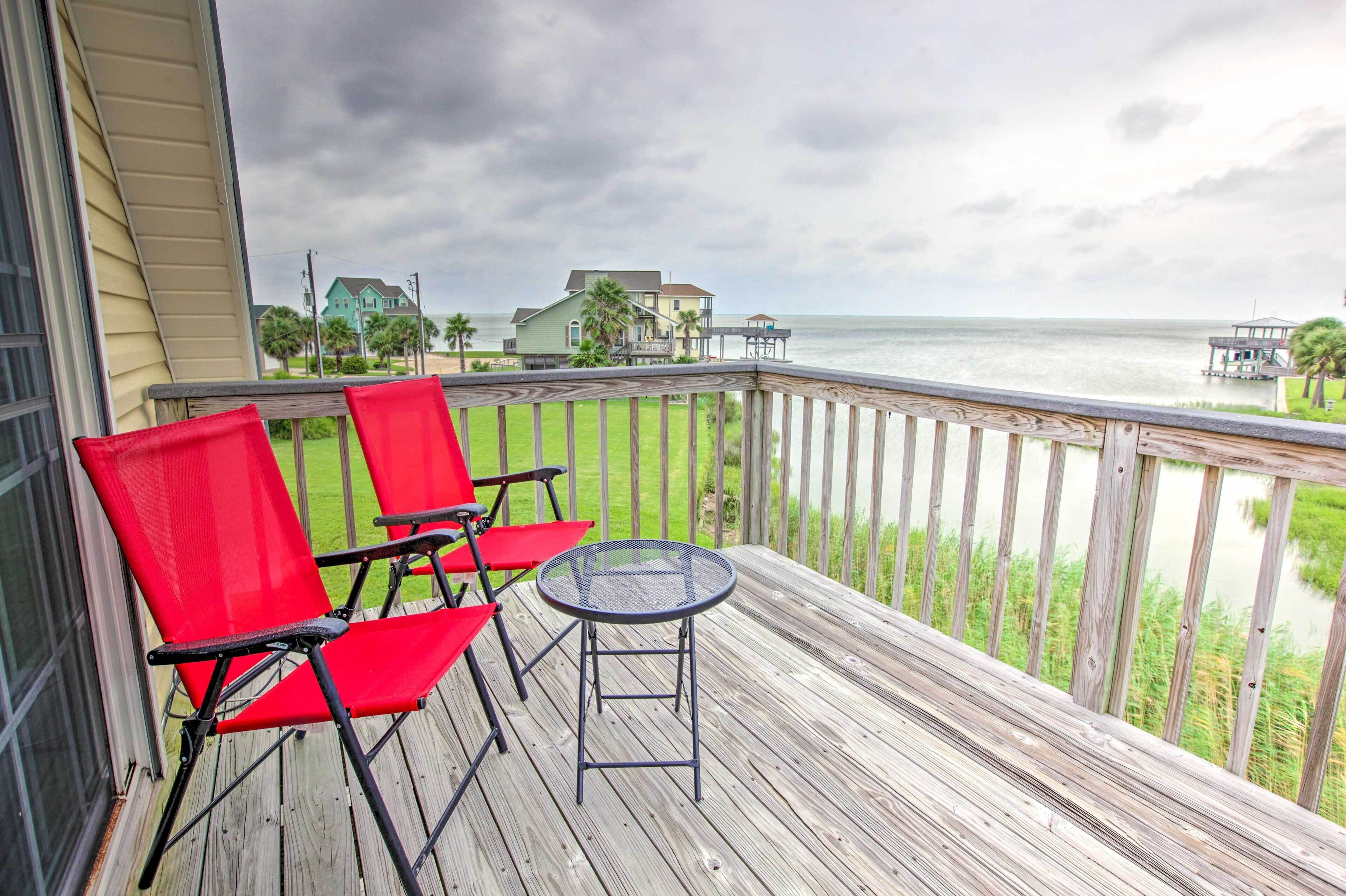A wooden deck with red chairs and a small table, overlooking a body of water and houses.