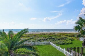 A coastal landscape with a white picket fence, green grass, and a view of the ocean. - Steps to Private Beach! Pet-friendly Getaway (Galveston)