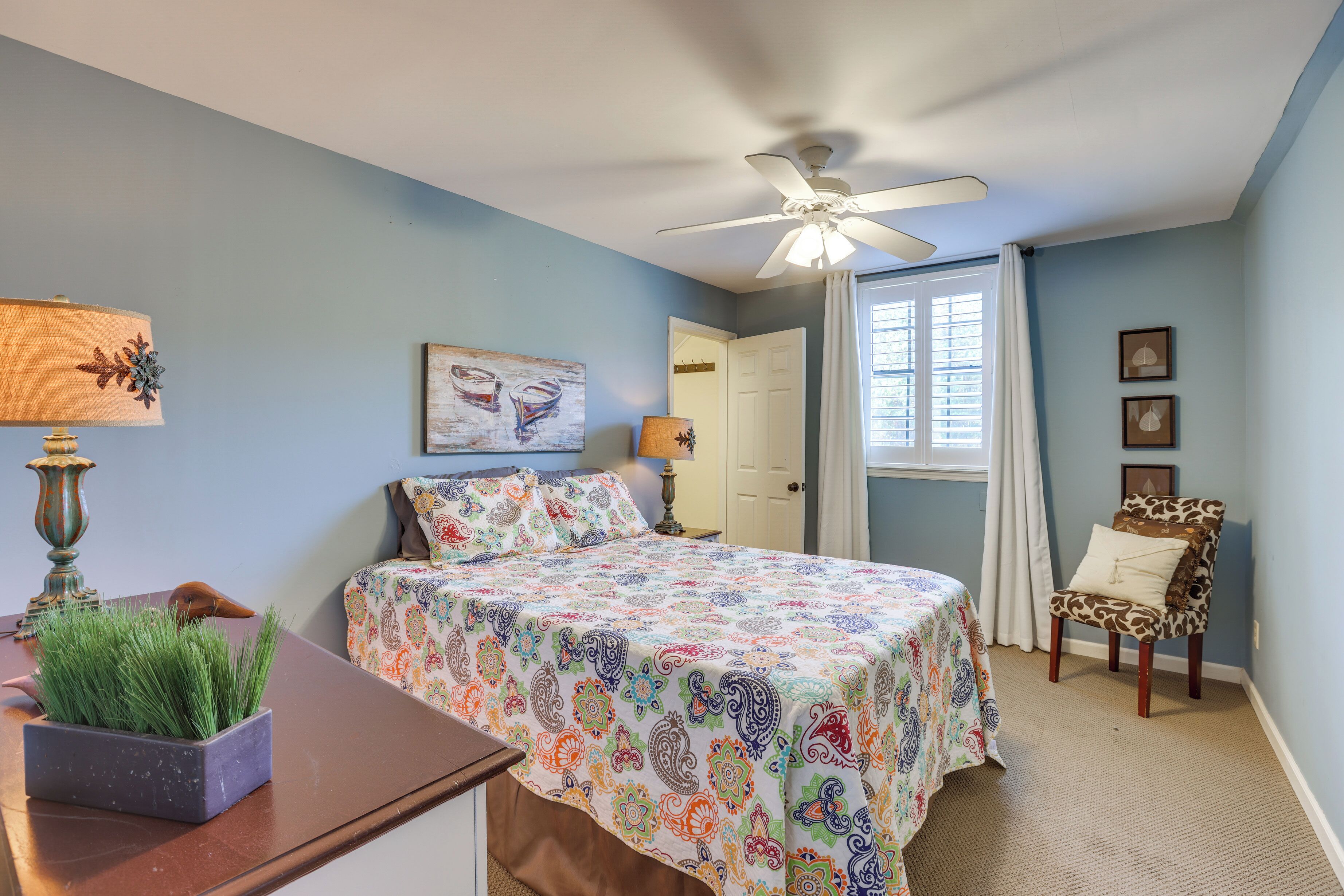 A bedroom with a bed, a chair, a desk with a lamp, and a ceiling fan.