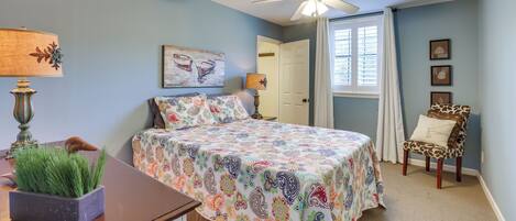 A bedroom with a bed, a chair, a desk with a lamp, and a ceiling fan.
