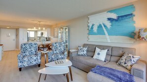 A living room with a sofa, armchairs, and a coffee table. There is a dining area with a table and chairs. A large painting of palm trees is on the wall. - Cozy Condo w/ Gulf View, Bird Watching + Telescope (Hudson)