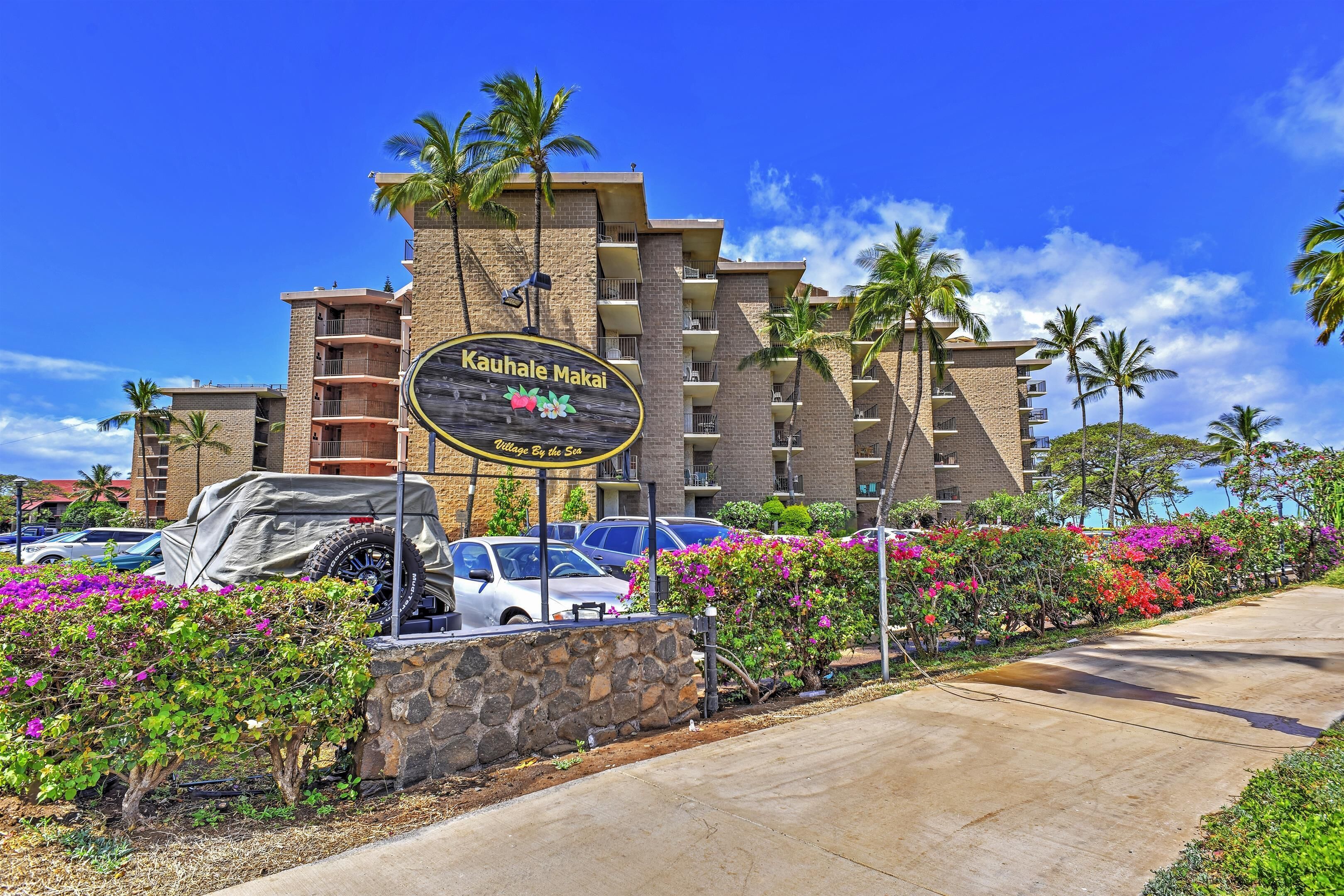 Hotel con un letrero que dice "Kauhale Makai" y vista al estacionamiento con coches y exuberante vegetación.