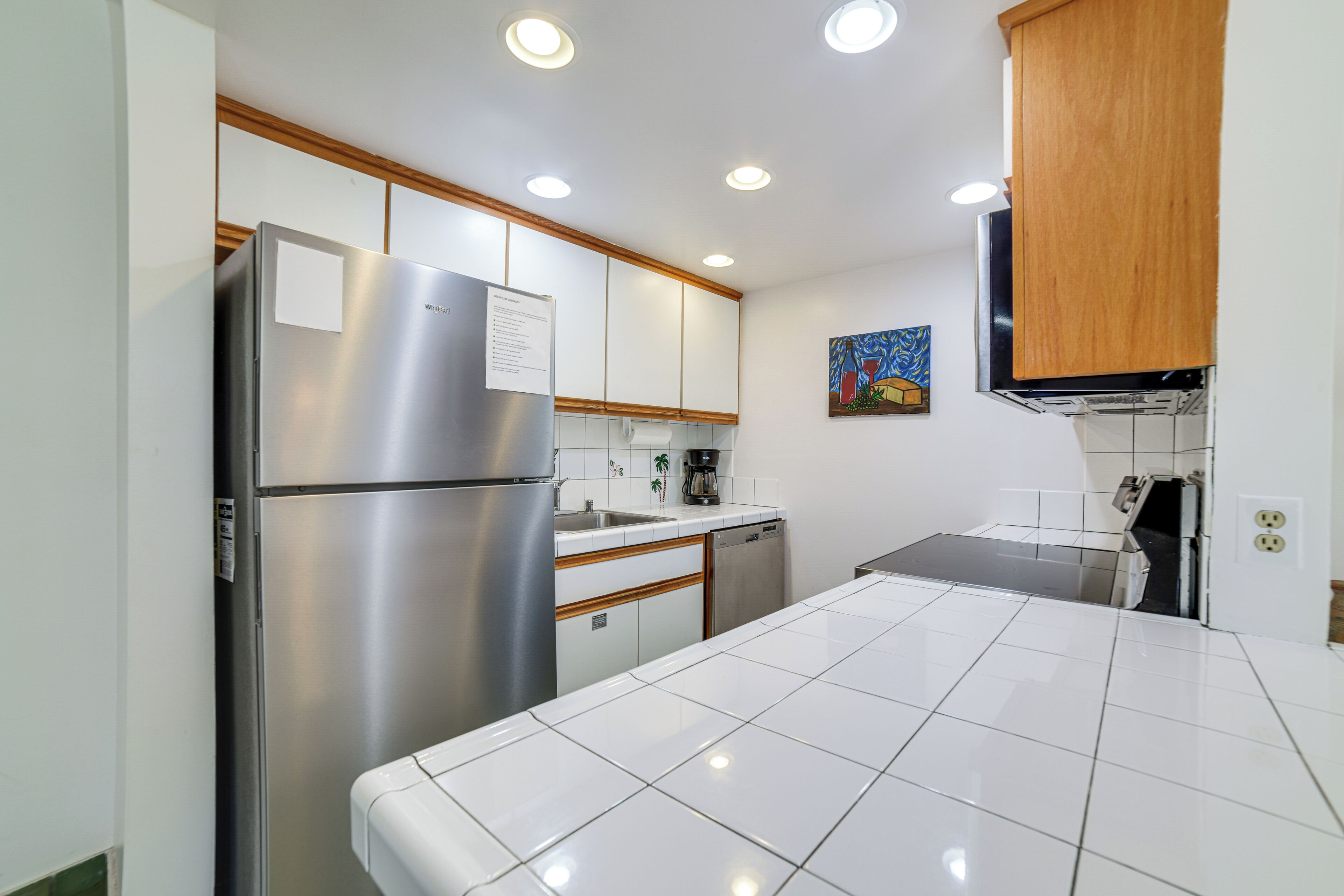 A modern kitchen with a stainless steel refrigerator, wooden cabinets, white tiles, and recessed ceiling lights.