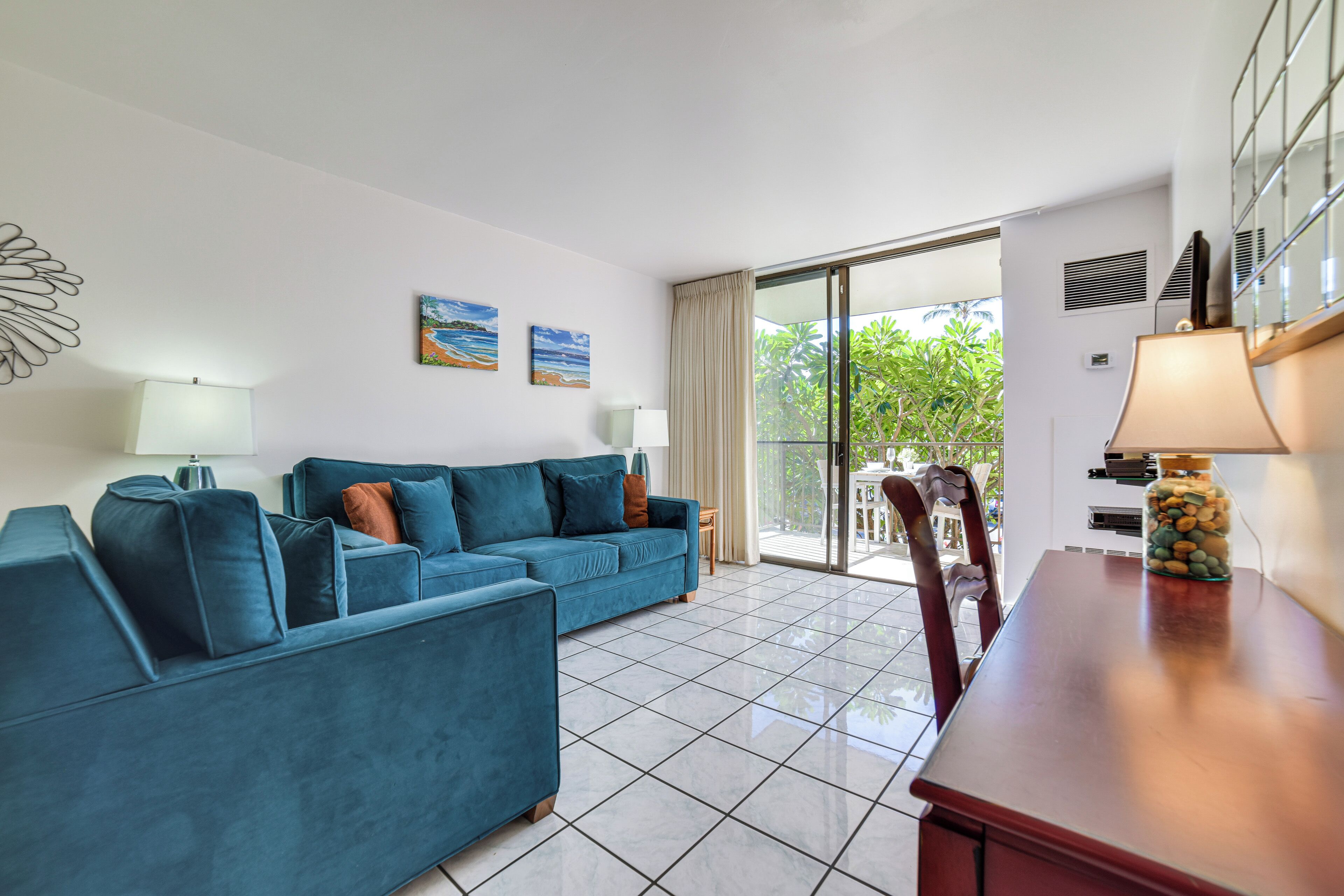 A modern living room with a blue sofa set, a wooden dining table, and a glass door leading to a balcony with a view of greenery.