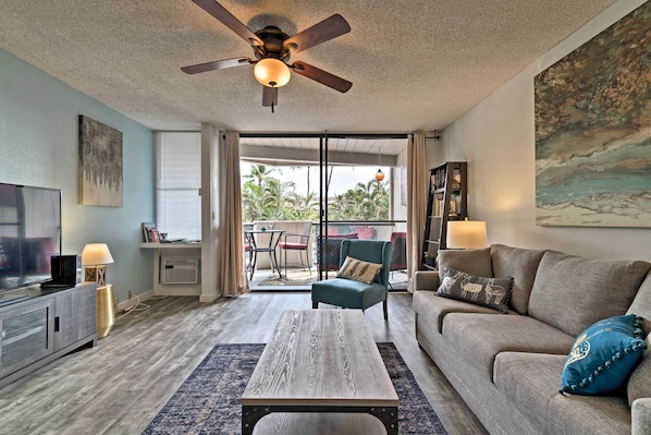 A living room with a ceiling fan, a TV, a sofa, a coffee table, and a view of palm trees through the sliding glass door. - Kailua-kona Condo w/ Lanai, Walk to Beach + Pier! (Kailua-Kona)