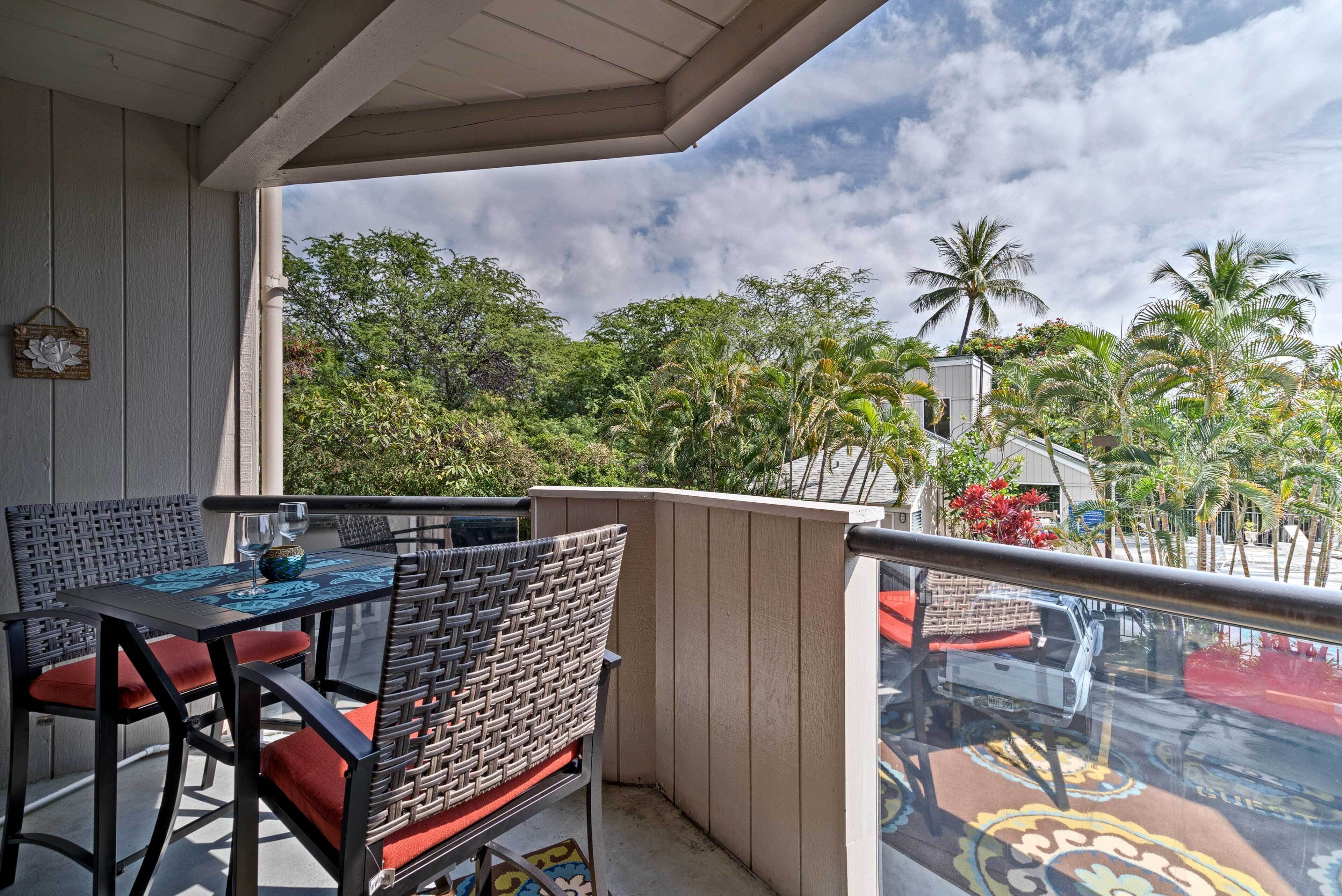 A balcony with a table and chairs, overlooking a lush garden with palm trees and a building in the background.