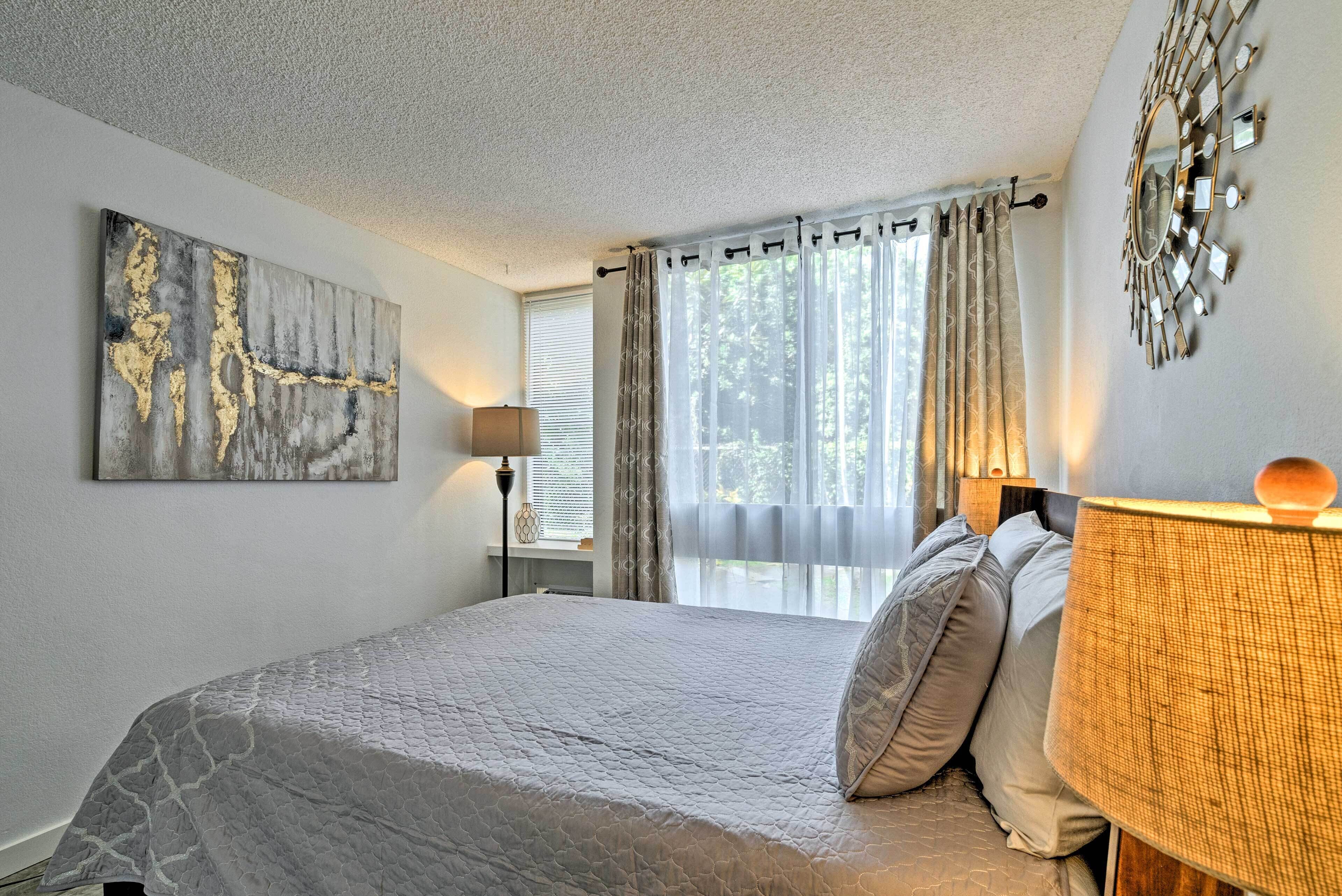 A bedroom with a bed, a lamp, a window with curtains, and a wall art piece.