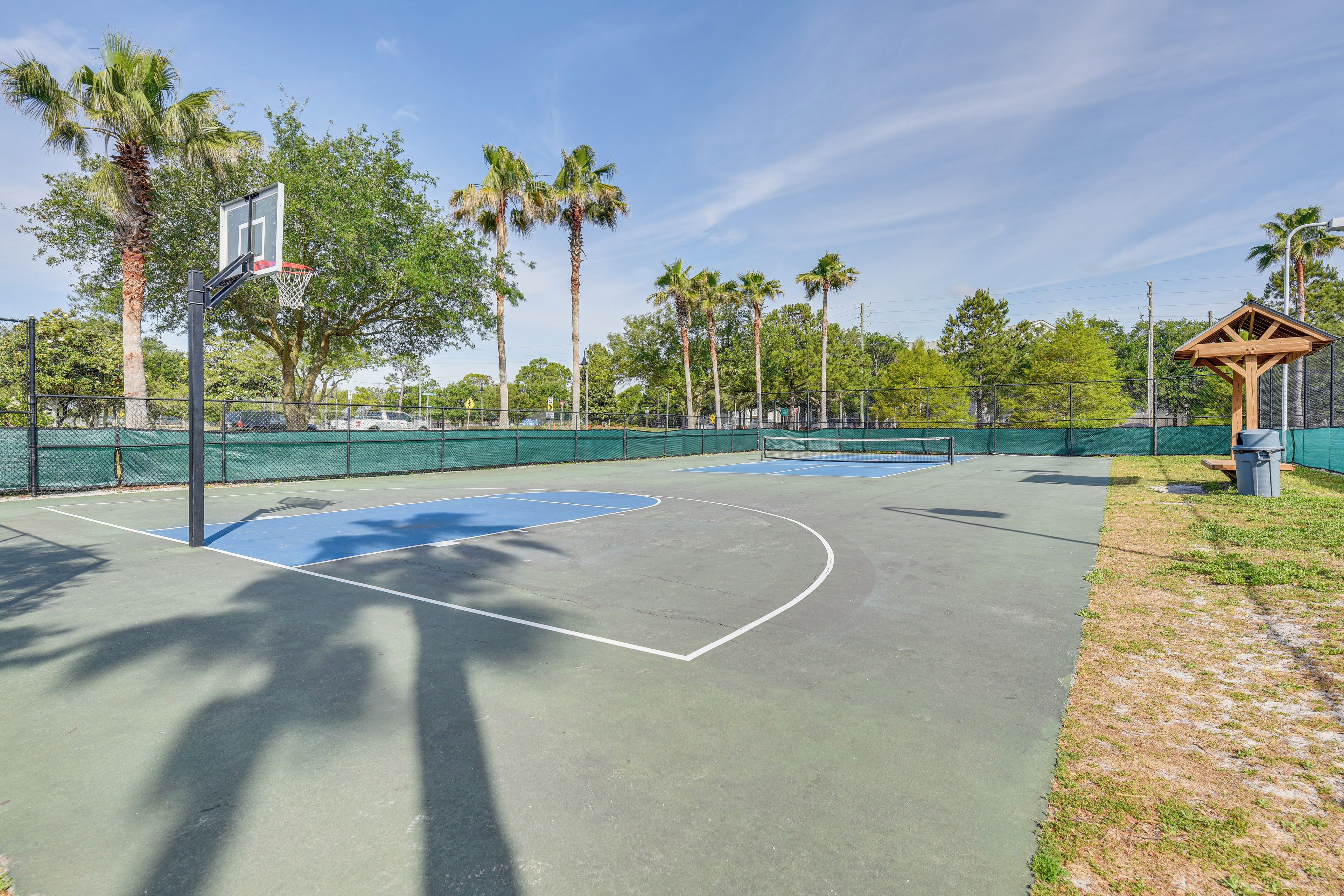 Een basketbalveld buiten met een houten paviljoen, omgeven door palmbomen en een groene hekken.