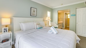 Een net opgemaakt bed met witte lakens en decoratieve kussens, een bedlamp, een ingelijst foto en een klein plankje met een boek en een afstandsbediening.