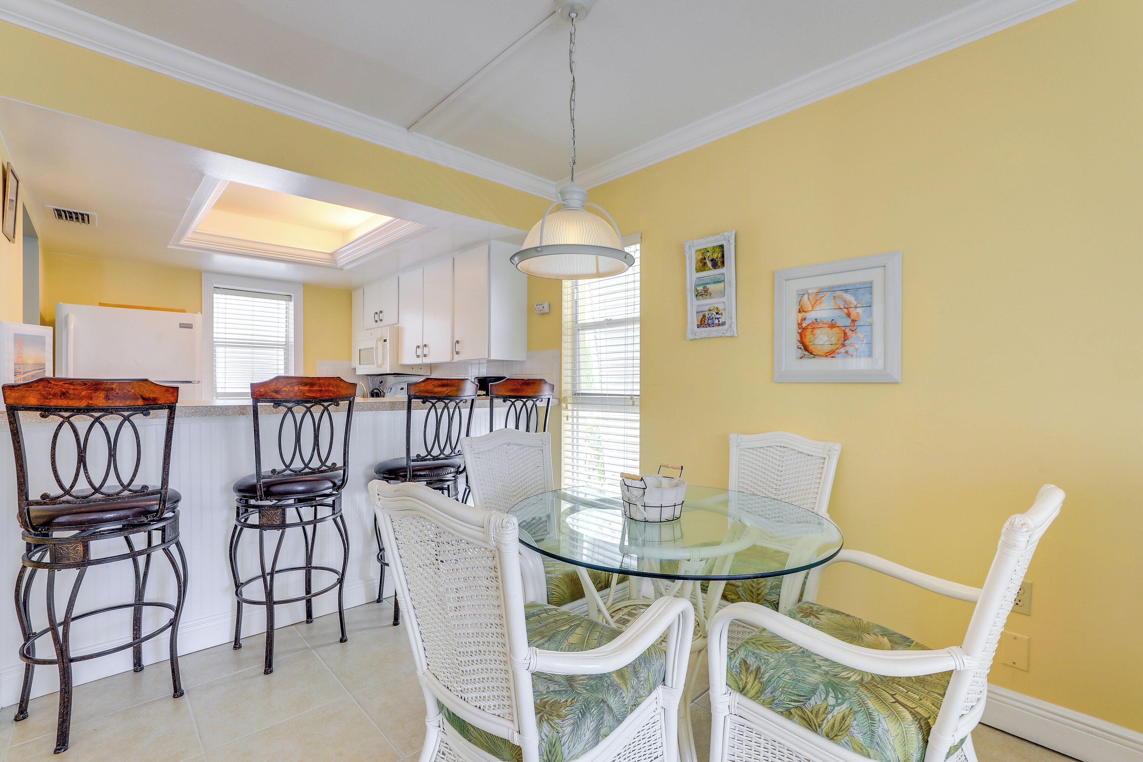 A dining area with a glass table and white wicker chairs, a kitchen in the background, and yellow walls.
