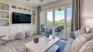 A modern living room with a flat-screen TV, white shelves, a ceiling fan, and a balcony with outdoor furniture.
