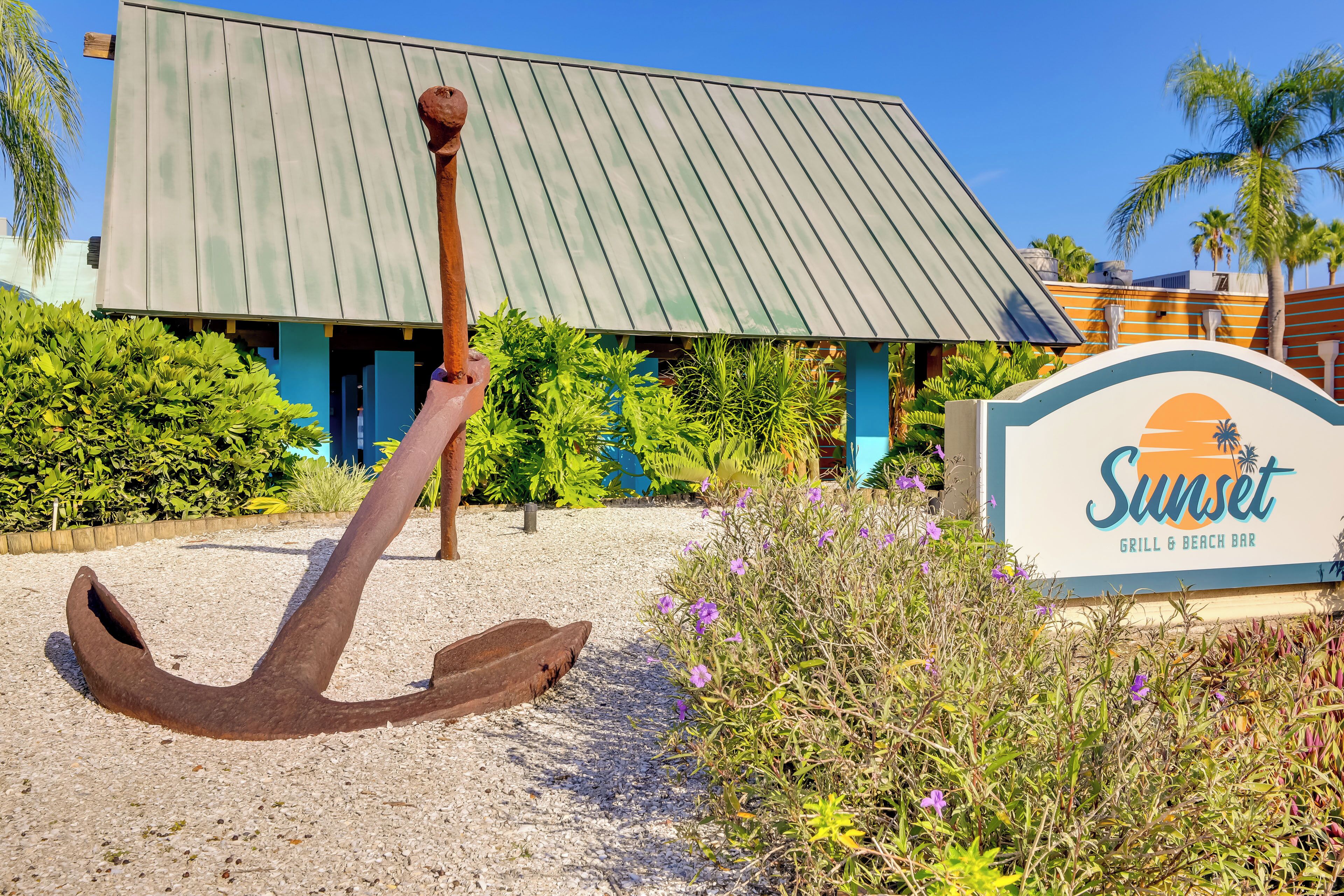 A building with a sign reading "Sunset Grill & Beach Bar" and an anchor sculpture in front.