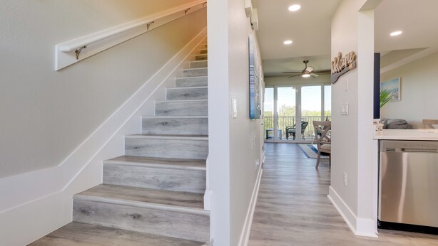 A modern kitchen with a stainless steel dishwasher, a wooden staircase, and a view into the living area.