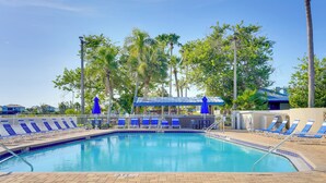 A swimming pool with blue lounge chairs and a covered area, surrounded by palm trees and a clear sky.