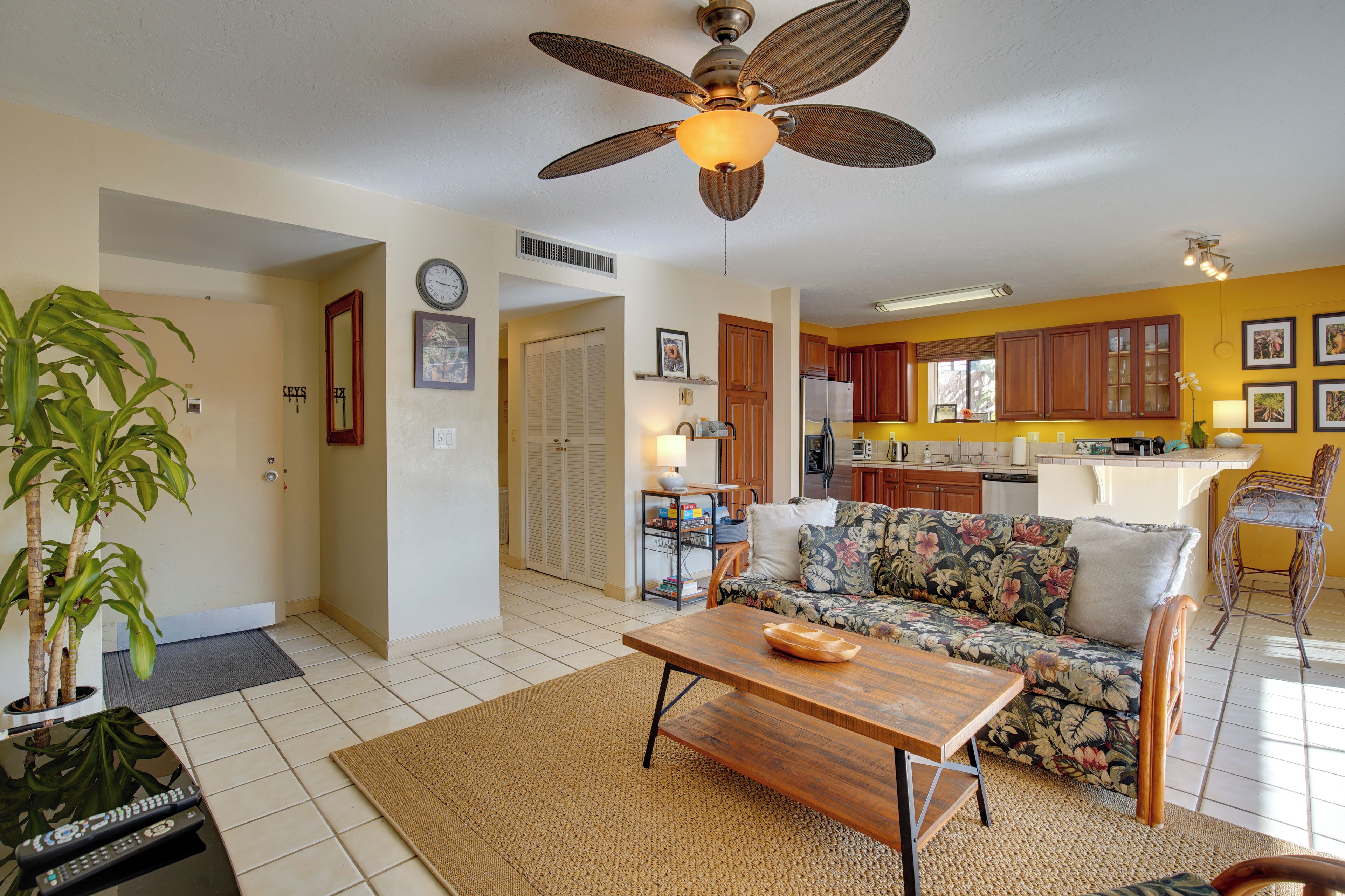 A living room with a sofa, coffee table, and a ceiling fan. There is a kitchen area with cabinets and a refrigerator.