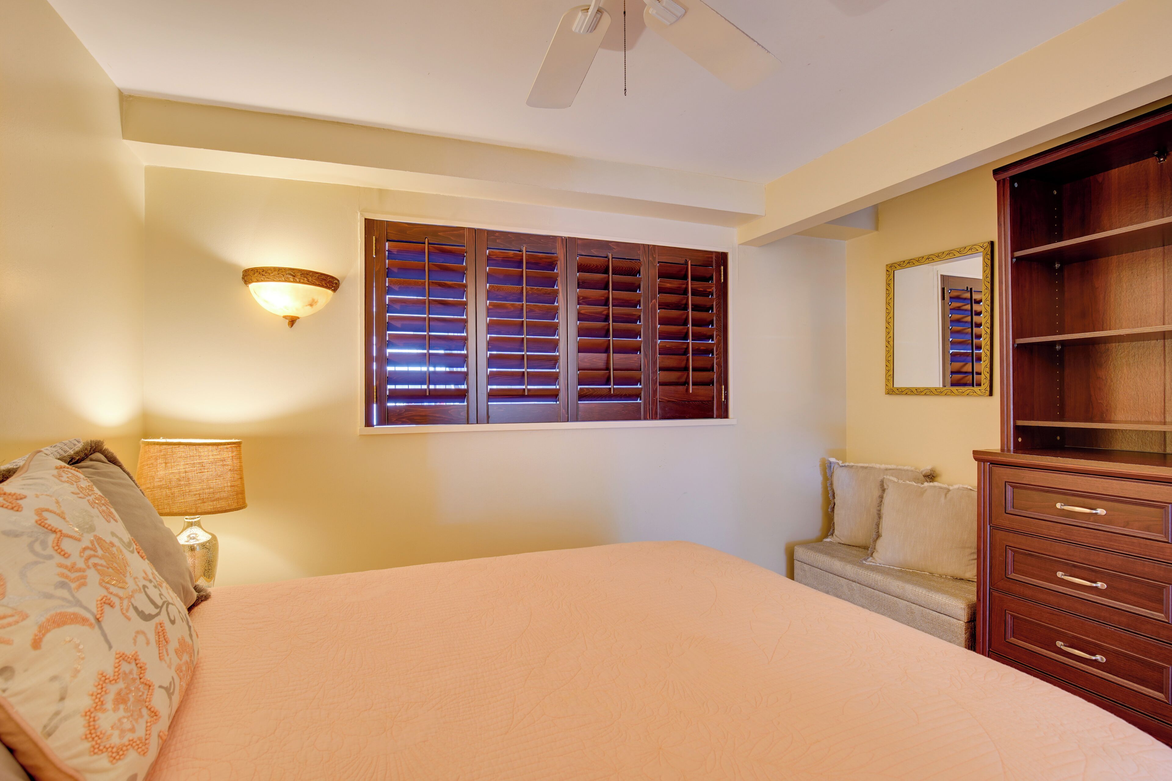 A bedroom with a bed, a dresser, a nightstand with a lamp, a window with blinds, and a ceiling fan.