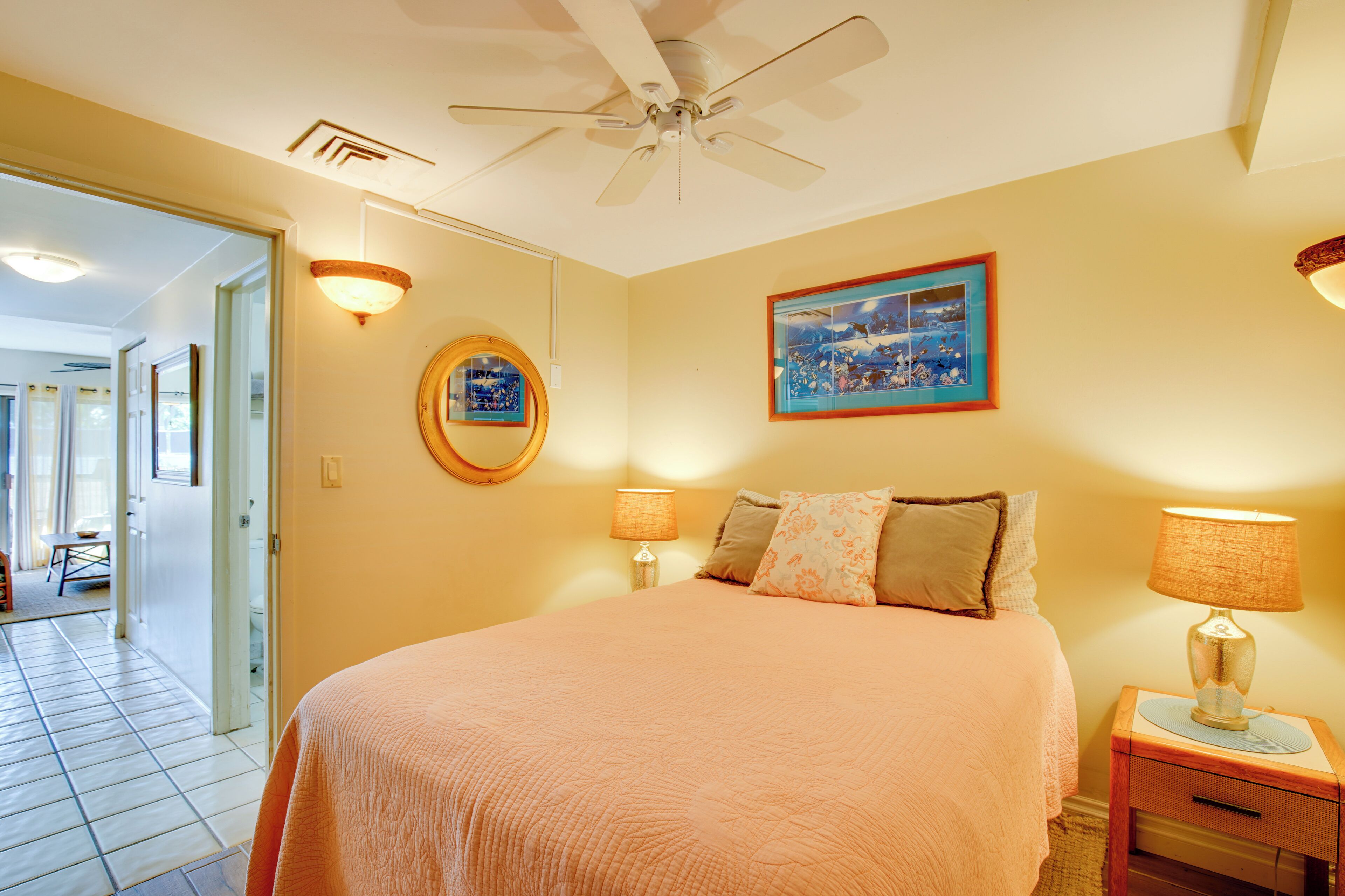 A bedroom with a bed, two bedside lamps, a ceiling fan, a framed picture, and a round mirror on the wall.