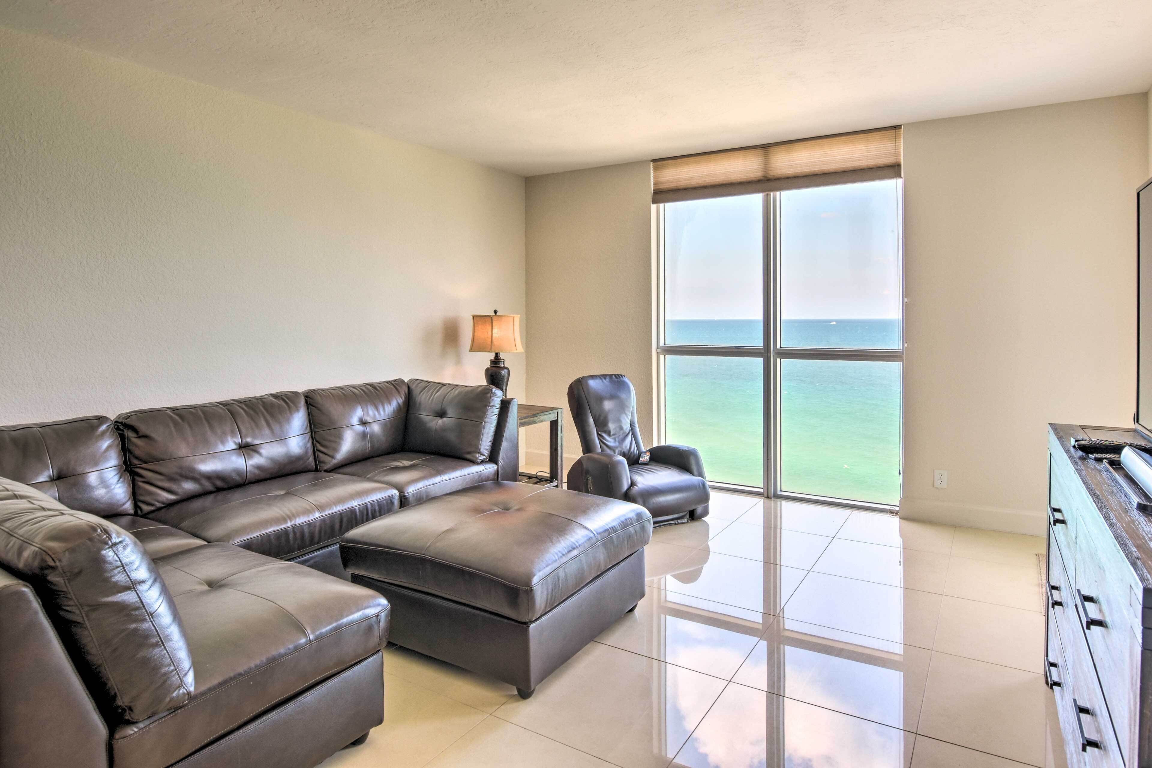 A living room with a sectional sofa, a chair, a lamp, and a view of the ocean through large windows.