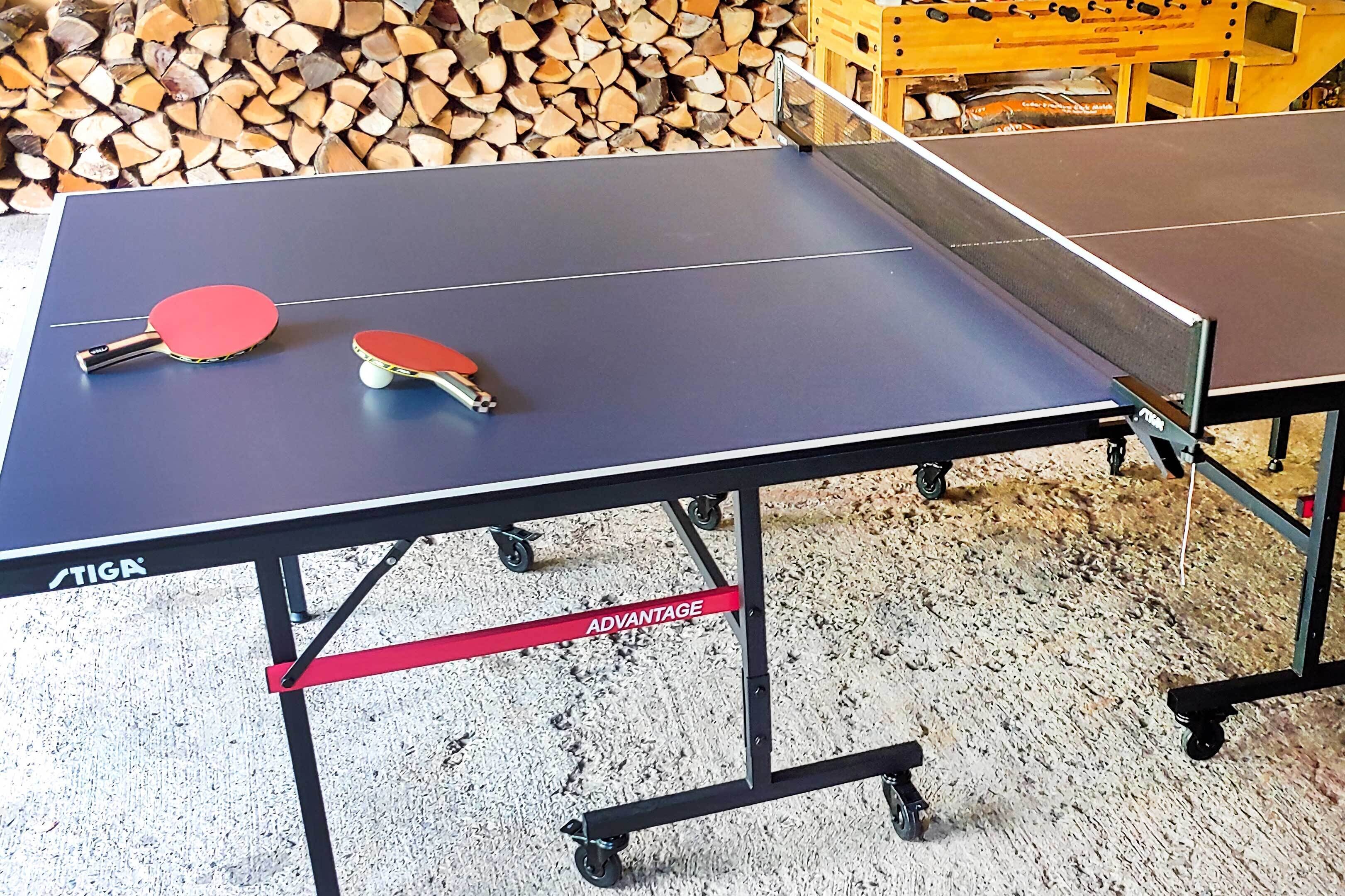 Une table de ping-pong avec deux raquettes et une balle de tennis de table, posée sur un sol où est fixé un ruban rouge portant l'inscription « AVANTAGE ».