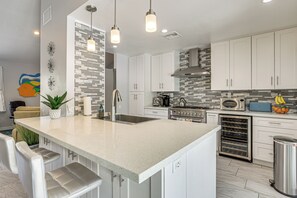 Une cuisine moderne avec des meubles blancs, un grand îlot central doté de tabourets, des appareils en acier inoxydable et un pare-feu orné de carreaux gris.