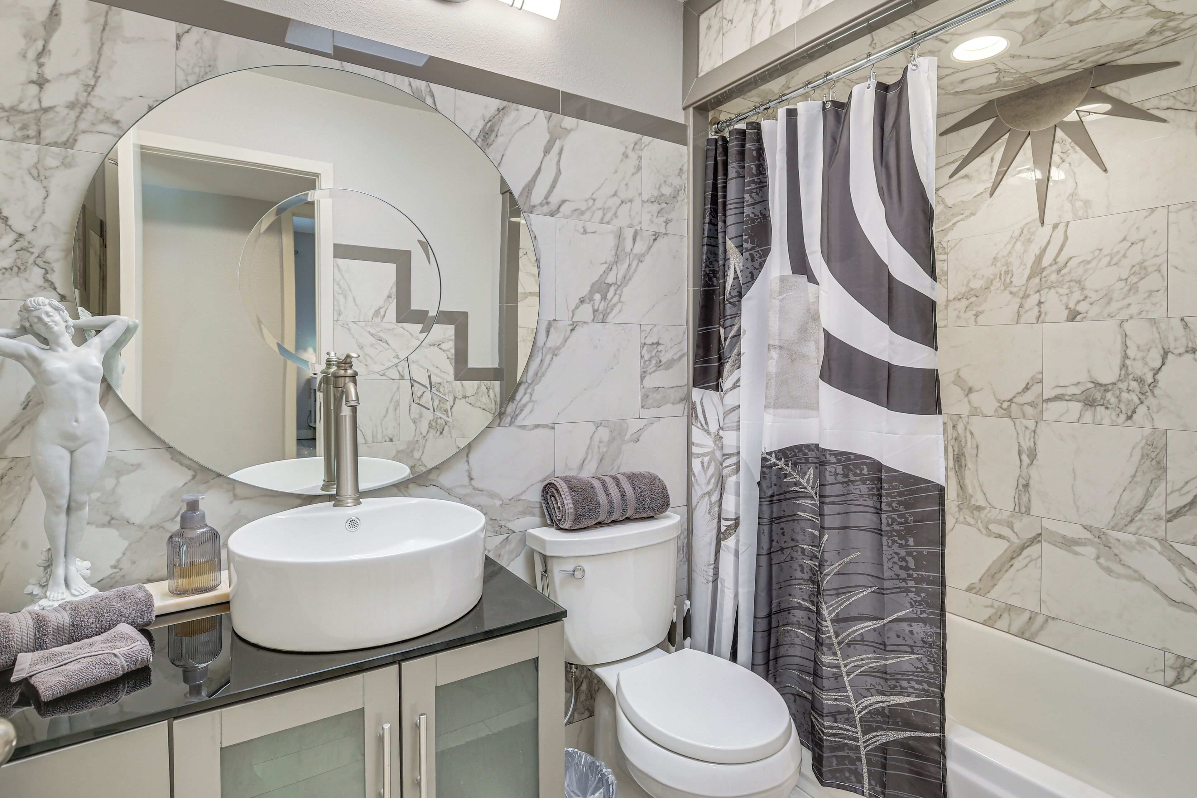 Une salle de bain avec un lavabo en marbre, des toilettes, une douche avec un rideau à motifs et une statue sur le côté gauche.